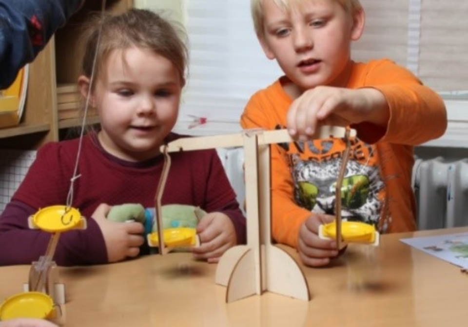 Das Bild zeigt zwei Kinder, die an einer Wippe experimentieren, um das Gleichgewicht zu pr&uuml;fen. Sie halten jeweils eine gelbe Schaufel in der Hand, w&auml;hrend sie gespannt beobachten, wie sich die Wippe bewegt.