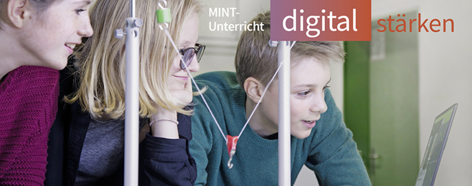 Das Bild zeigt drei Sch&uuml;ler, die konzentriert an einem Experiment oder einer digitalen Pr&auml;sentation arbeiten. Im Hintergrund sind Apparaturen sichtbar, die in einem MINT-Unterricht eingesetzt werden, um das Interesse an Naturwissenschaften zu f&ouml;rdern.