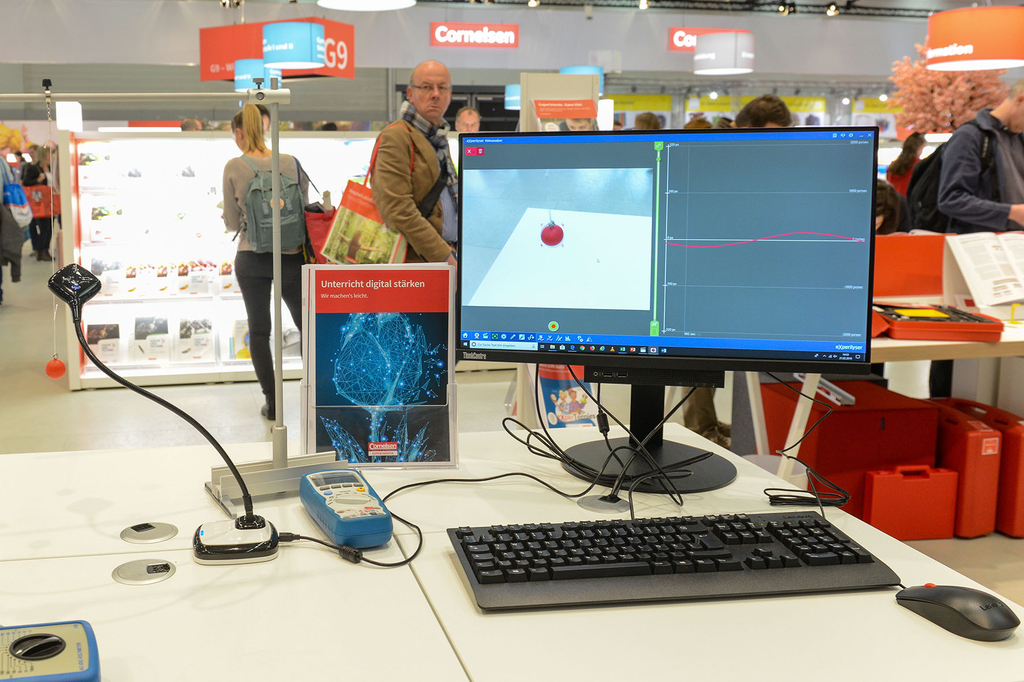 Das Bild zeigt einen Arbeitsplatz mit einem Computer, der ein Experiment auf einem Tisch dokumentiert, wo eine rote Kugel an einem Gerät hängt. Neben dem Monitor sind verschiedene wissenschaftliche Geräte und ein Informationsblatt platziert, das den digit