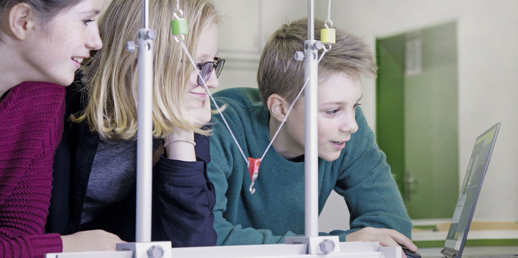 Das Bild zeigt drei Schüler, die aufmerksam an einem Experiment arbeiten, während sie einen Bildschirm beobachten. Vor ihnen befindet sich eine Versuchsanordnung mit Schnüren und Gewichten, die typische Materialien für den Experimentierbedarf darstellen.