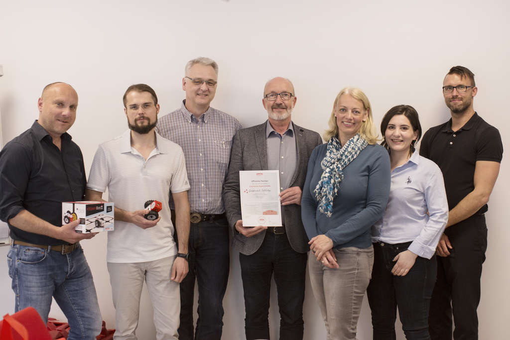 Das Bild zeigt eine Gruppe von sechs Personen, die zusammen stehen und verschiedene Produkte sowie ein Zertifikat in der Hand halten. Sie scheinen an einem Projekt im Bereich Experimentierbedarf für das Bildungswesen beteiligt zu sein.