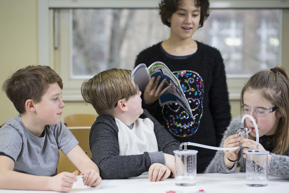 Das Bild zeigt eine Gruppe von Kindern, die an einem Experiment arbeiten. Ein M&auml;dchen erkl&auml;rt etwas aus einem Buch, w&auml;hrend ein anderes Kind mit einem wissenschaftlichen Ger&auml;t in zwei Bechern experimentiert.