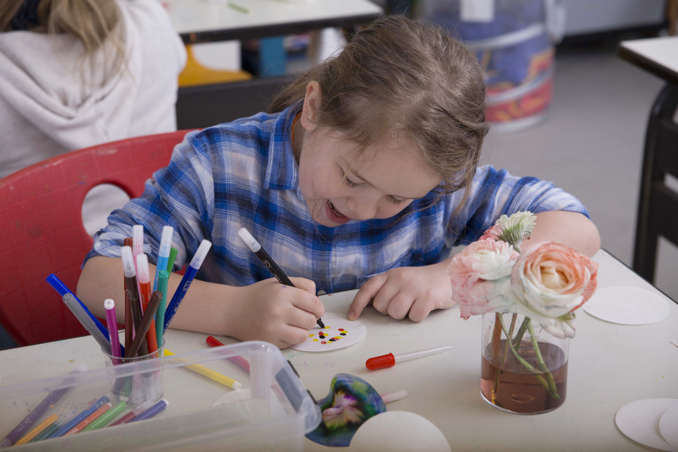 Das Bild zeigt ein M&auml;dchen, das konzentriert mit Stiften auf einer kleinen wei&szlig;en Fl&auml;che malt. Auf dem Tisch befinden sich verschiedene Marker, eine Pipette und eine Vase mit Blumen.