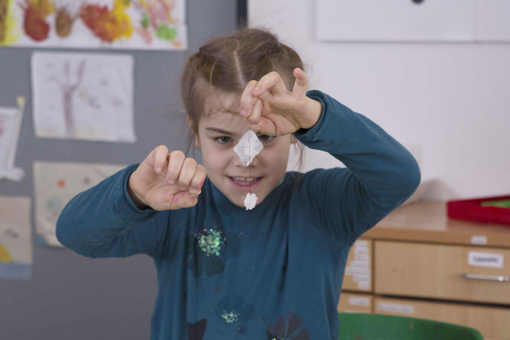 Ein Mädchen hält stolz ein kleines Experiment in der Hand, das aus einem transparenten, gefalteten Material und etwas Weißem besteht. Im Hintergrund sind Zeichnungen von anderen Kindern zu sehen, die eine kreative Umgebung schaffen.