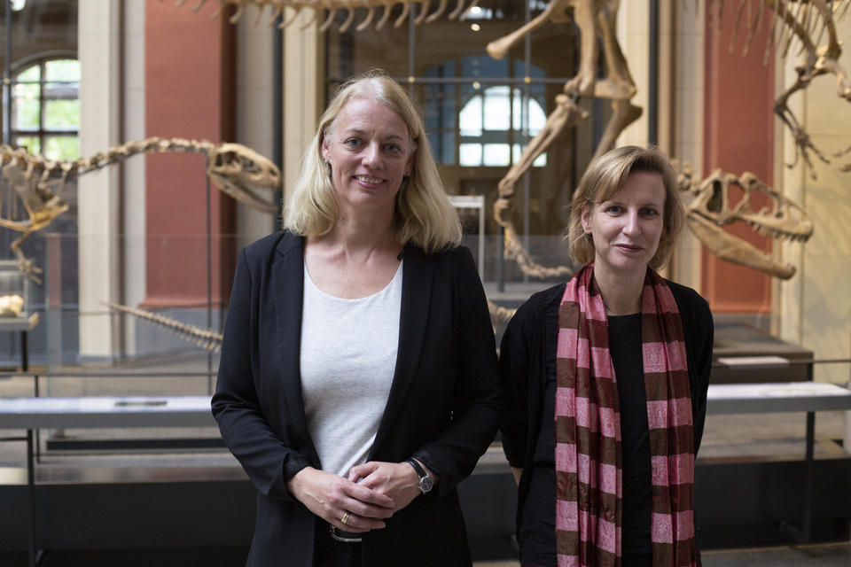 Das Bild zeigt zwei Frauen, die l&auml;chelnd in einem Museum stehen, in dem Dinosaurier-Skelette im Hintergrund sichtbar sind. Der Raum scheint hell und architektonisch eindrucksvoll zu sein, was die Bedeutung des Bildungsangebots unterstreicht.