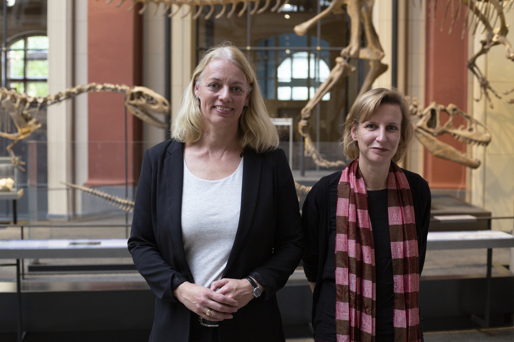 Das Bild zeigt zwei Frauen, die lächelnd in einem Museum stehen, in dem Dinosaurier-Skelette im Hintergrund sichtbar sind. Der Raum scheint hell und architektonisch eindrucksvoll zu sein, was die Bedeutung des Bildungsangebots unterstreicht.