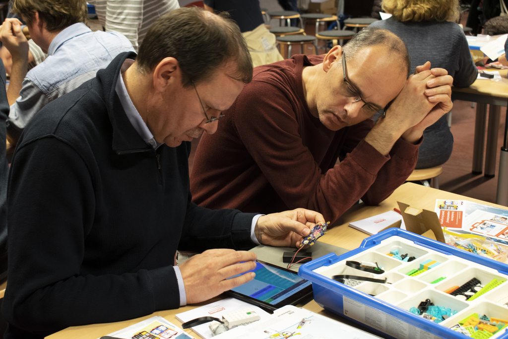 Das Bild zeigt zwei Männer, die konzentriert an einem Tisch arbeiten, während sie ein elektronisches Bauteil und ein Tablet untersuchen. Auf dem Tisch liegen verschiedene Materialien und Werkzeuge für Experimente, die für das Bildungswesen gedacht sind.