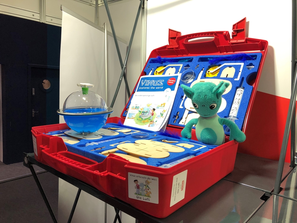 Das Bild zeigt eine rote Kunststoffkiste, die mit Experimentiermaterialien f&uuml;r den Unterricht ausgestattet ist. Darin befinden sich ein Wasserbeh&auml;lter, verschiedene Werkzeuge und ein gr&uuml;nes Stofftier, das als Maskottchen dient.
