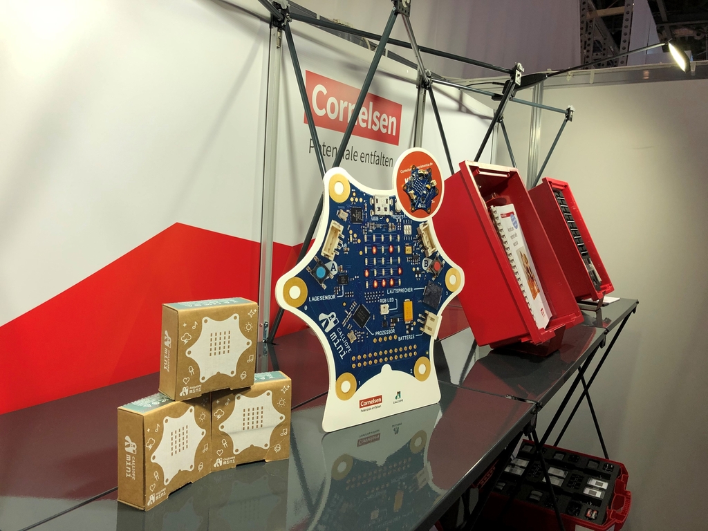 Das Bild zeigt einen Messestand mit verschiedenen Lehrmaterialien f&uuml;r das deutsche Bildungswesen. Im Vordergrund ist eine Schaltung zu sehen, umgeben von gestapelten Boxen mit zus&auml;tzlichen Ger&auml;ten oder Modulen.