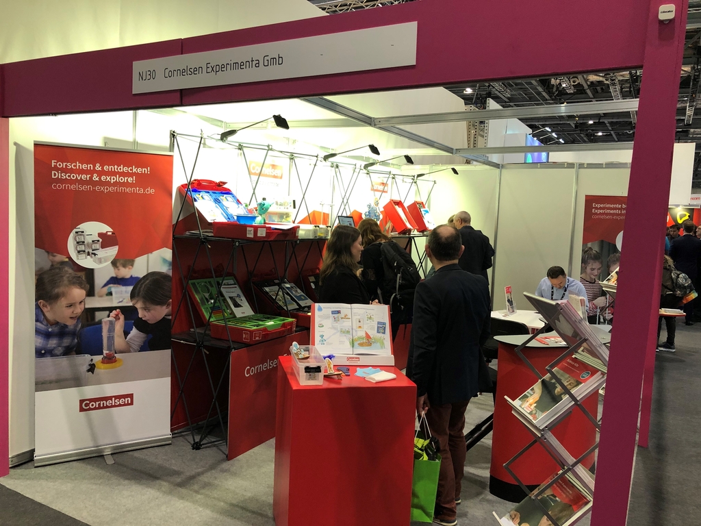 Das Bild zeigt den Stand der Cornelsen Experimenta GmbH auf einer Bildungsmesse, wo verschiedene Experimentiermaterialien und Bildungsunterlagen ausgestellt sind. Besucher interagieren mit den Produkten und Informationen.