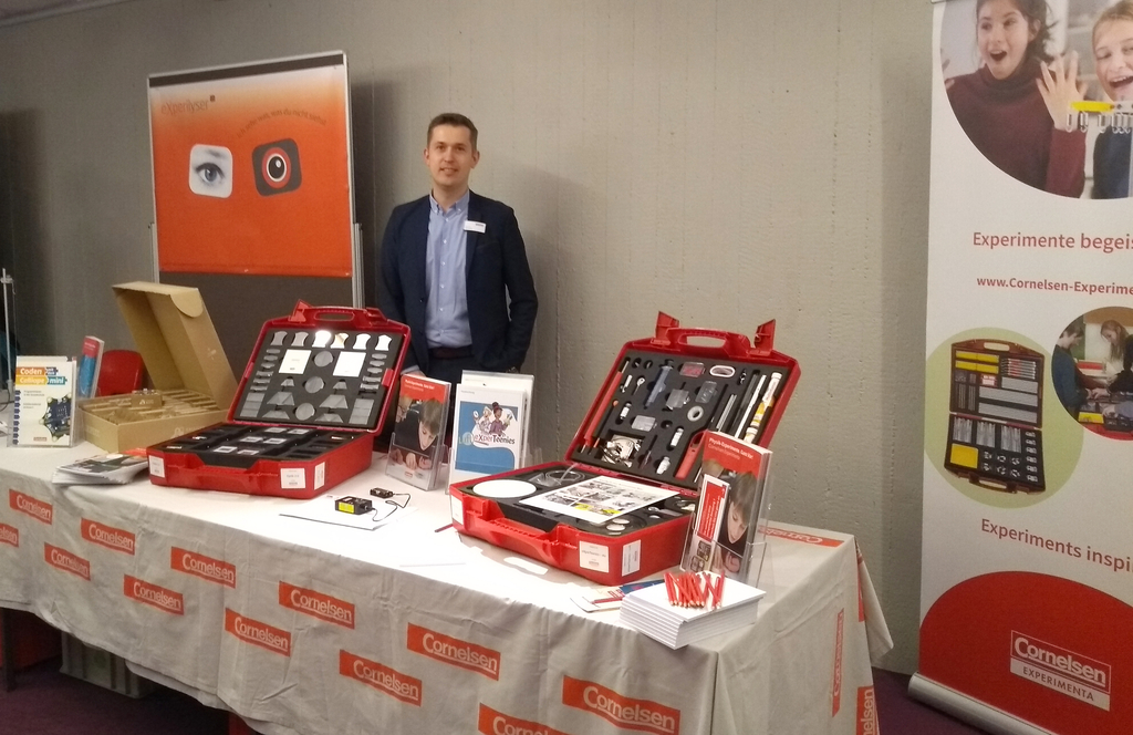 Das Bild zeigt einen Stand mit Experimentiermaterialien f&uuml;r das Bildungswesen, einschlie&szlig;lich zwei roter Koffer mit verschiedenen Experimentierwerkzeugen. Im Hintergrund steht ein Mann, der den Stand pr&auml;sentiert.