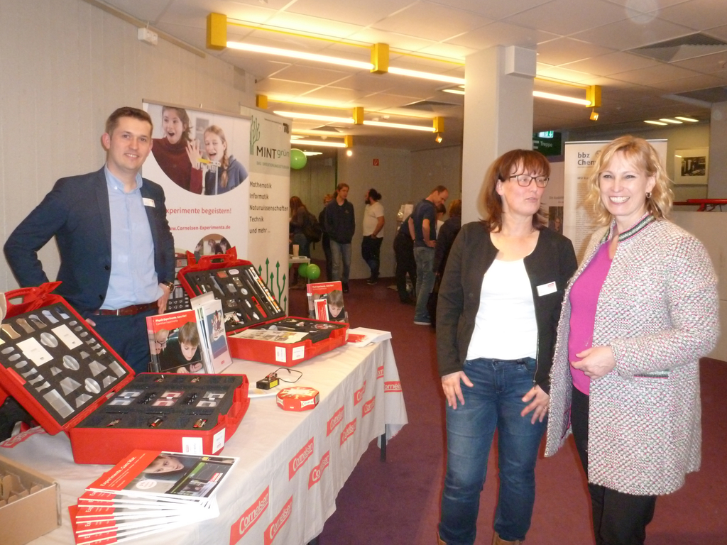 Das Bild zeigt einen Stand für Experimentierbedarf im Bildungsbereich, mit mehreren roten Experimentierkoffern, die verschiedene Experimente und Materialien enthalten. Drei Personen stehen im Vordergrund und unterhalten sich, während im Hintergrund weiter