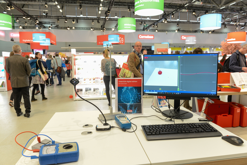 Das Bild zeigt einen Messplatz mit einem Computer, auf dem eine Versuchsanordnung mit einer roten Kugel zu sehen ist. Im Hintergrund sind Besucher auf einer Messe für Experimentierbedarf zu erkennen, umgeben von Informationsständen und Angeboten für das B