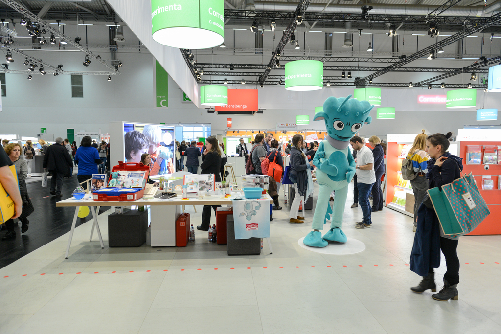 Das Bild zeigt einen Messestand mit einem gro&szlig;en blauen Maskottchen und mehreren Personen, die sich um St&auml;nde mit Experimentiermaterialien versammeln. Die Umgebung ist hell und modern, mit verschiedenen Displays und Informationsst&auml;nden.