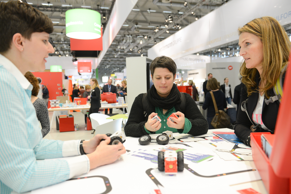 Das Bild zeigt drei Frauen, die an einem Tisch mit Experimentiermaterialien und arbeiten, darunter ist auch der Lernroboter eXperiBot. Im Hintergrund sind St&auml;nde und Besucher einer Messe zu sehen, was auf eine bildungsorientierte Veranstaltung hinweist.