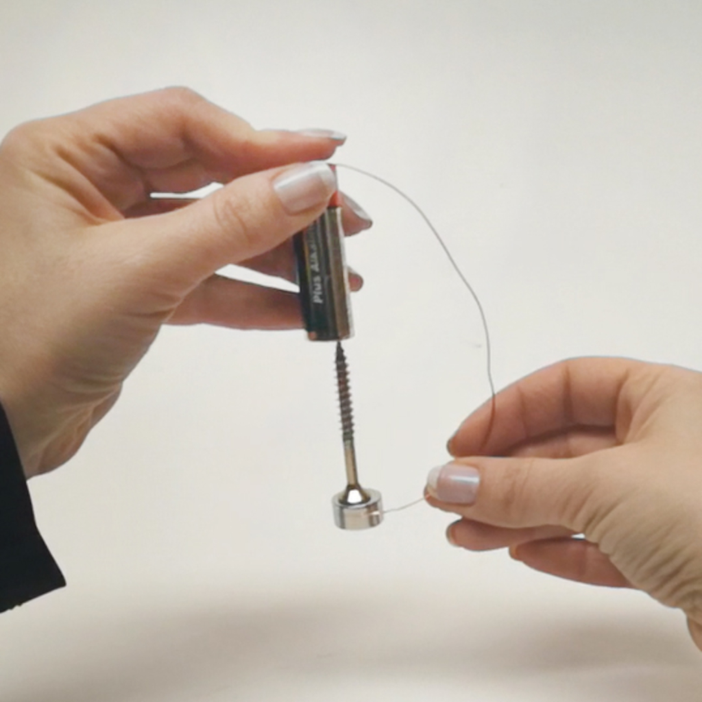 Das Bild zeigt zwei Hände, die eine Batterie und einen Draht halten. Der Draht ist an einem metallischen Objekt befestigt, das wahrscheinlich Teil eines Experimentes zur Demonstration von elektrischer Energie oder Magnetismus ist.