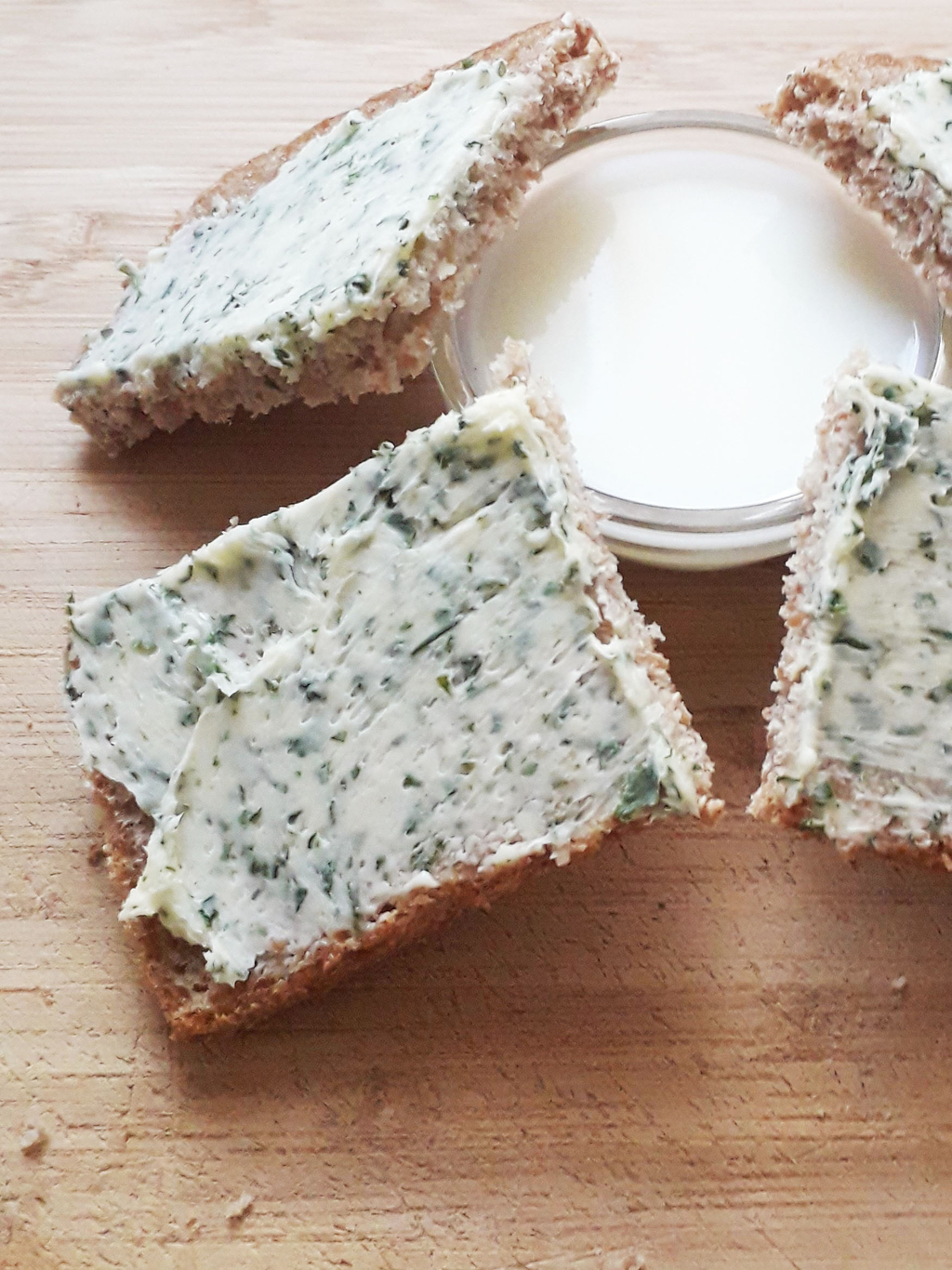 Das Bild zeigt vier Scheiben Vollkornbrot, die mit einer Kräuterbutter bestrichen sind. In der Mitte des Bildes befindet sich ein kleines Glas mit einer cremigen, weißen Substanz, wahrscheinlich einer weiteren Art von Aufstrich oder Dip.