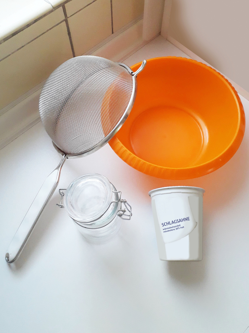 Das Bild zeigt eine orangefarbene Sch&uuml;ssel, ein feinmaschiges Sieb, ein rundes Glasgef&auml;&szlig; mit Verschluss und einen Becher mit der Aufschrift &bdquo;Schlagsahne&ldquo;. Dies sind Materialien und Bestandteile, um Butter herzustellen.