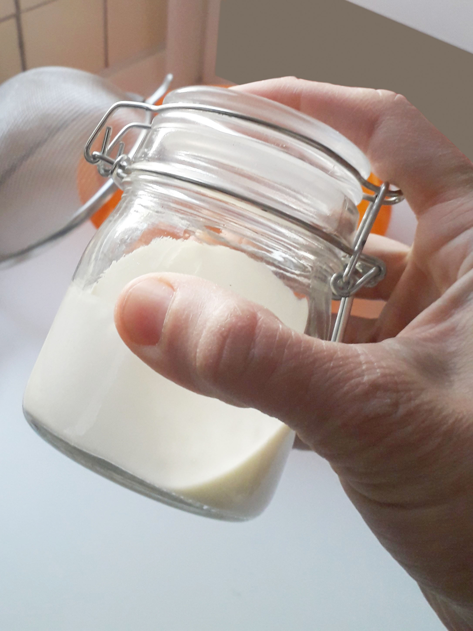 Das Bild zeigt eine Hand, die ein Glas mit Sahne h&auml;lt. Im Hintergrund ist ein Sieb zu sehen, das auf ein Experiment hindeutet wie die Herstellung von Buttermilch oder Butter.