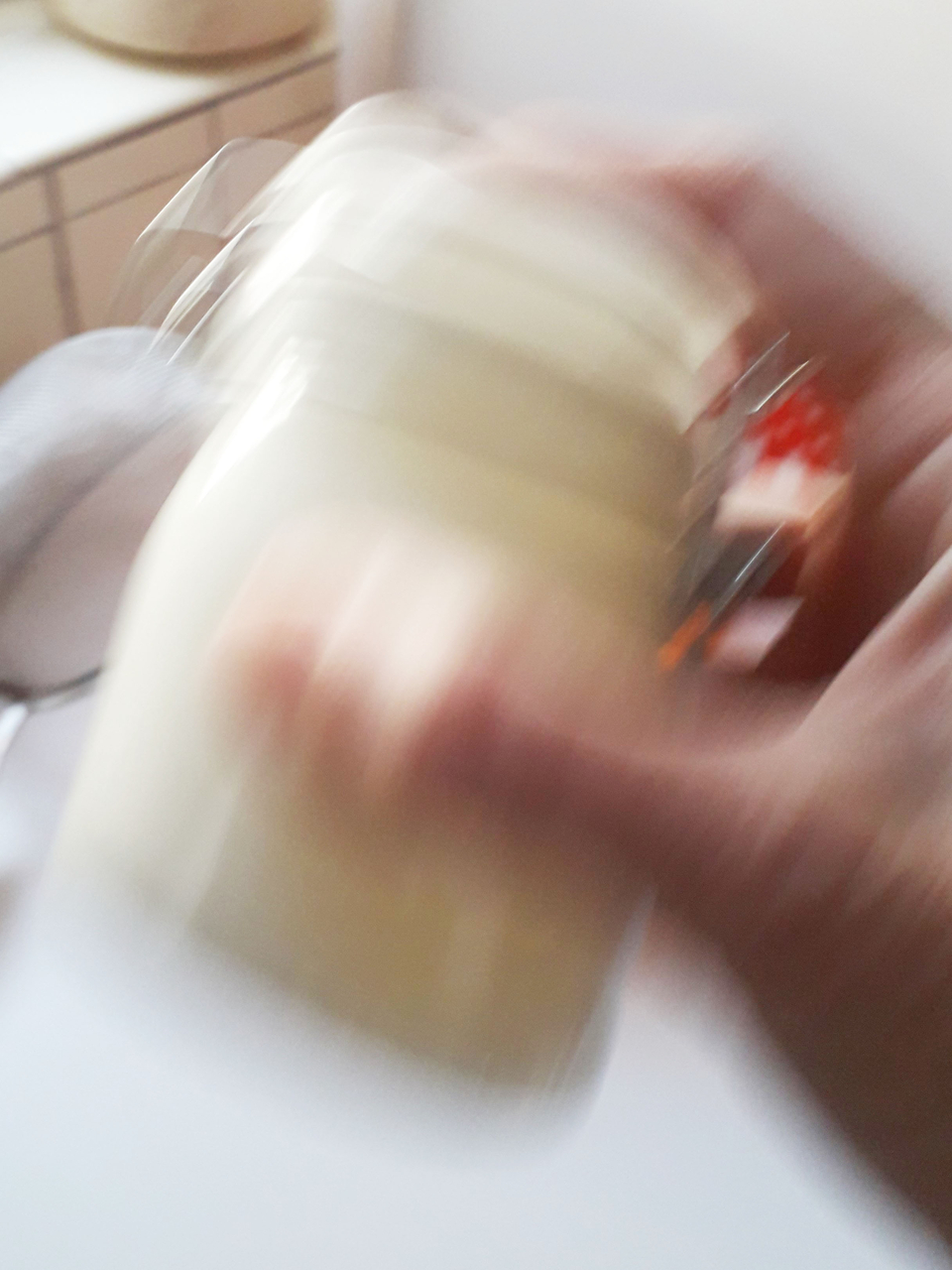 Das Bild zeigt eine verschwommene Hand, die ein Glas h&auml;lt, w&auml;hrend es gesch&uuml;ttelt wird. Im Hintergrund sind unscharfe K&uuml;chenelemente zu erkennen, was auf eine Experimentier- oder Kochsituation hindeutet.