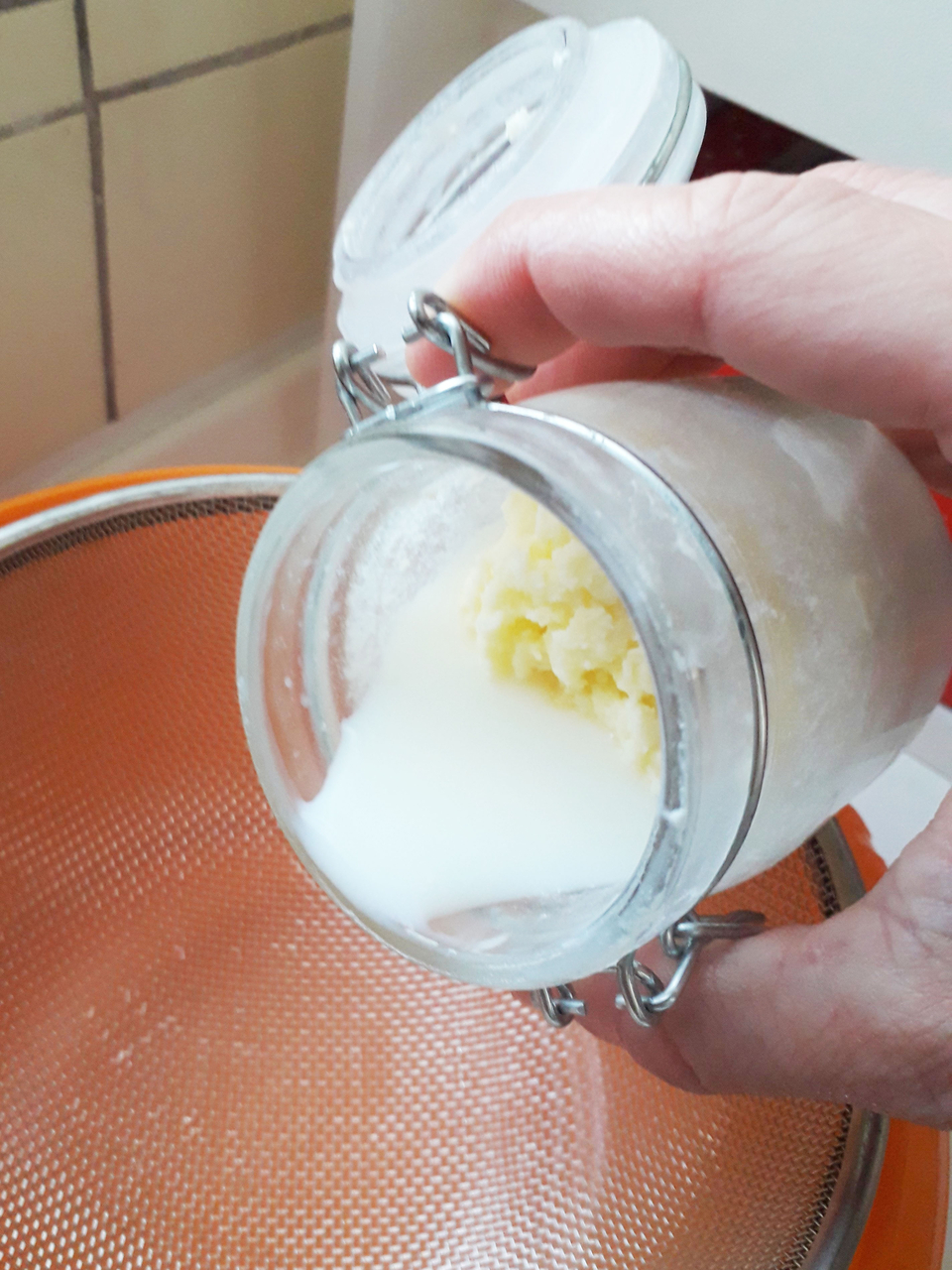 Das Bild zeigt ein Glas, das eine Mischung aus einer festen und einer fl&uuml;ssigen Substanz enth&auml;lt, w&auml;hrend es &uuml;ber einem Sieb gehalten wird. Der Kontext deutet auf einen Experimentierprozess hin, m&ouml;glicherweise im Bereich Chemie oder Biologie f&uuml;r den Bildu