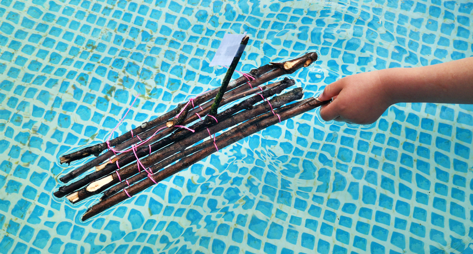 Das Bild zeigt ein Miniatur-Flo&szlig;, das aus mehreren zusammengebundenen &Auml;sten besteht und auf der Wasseroberfl&auml;che schwimmt. Eine Hand h&auml;lt das Boot, w&auml;hrend es in einem blauen Wasserbecken bleibt.