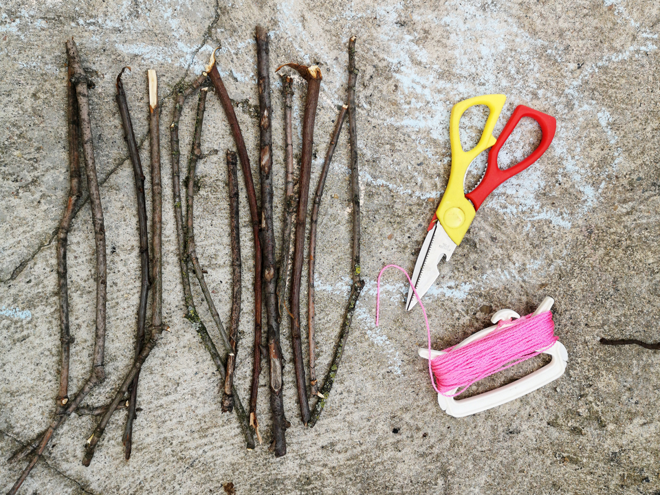 Das Bild zeigt eine Anordnung von mehreren d&uuml;nnen &Auml;sten auf einem grauen Untergrund, neben denen eine bunte Schere sowie ein Spulenhalter mit pinkem Faden liegen. Diese Materialien sind ideal f&uuml;r verschiedene kreative Experimente in der Bildung.
