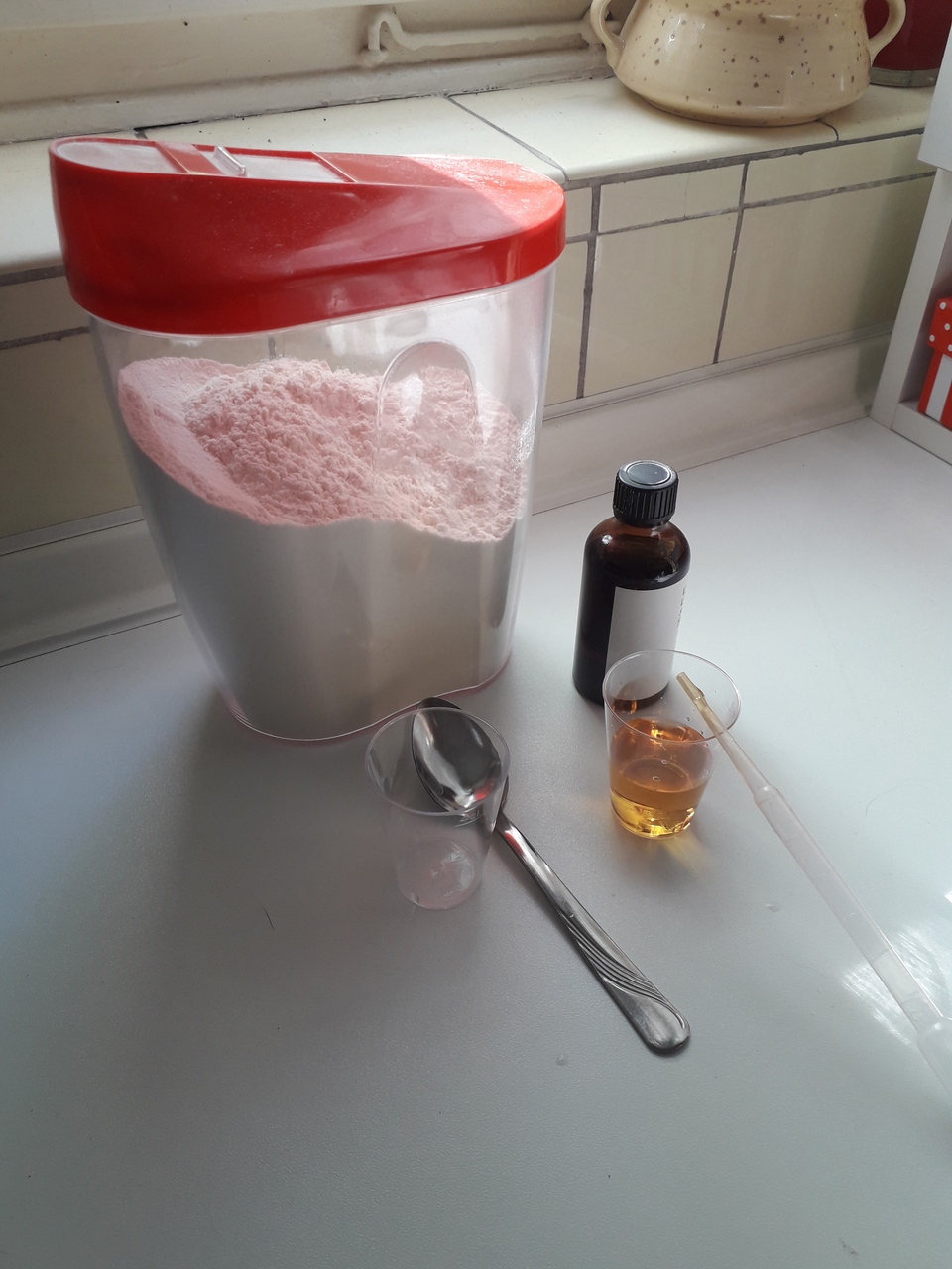 Das Bild zeigt einen gro&szlig;en Beh&auml;lter mit pinkem Pulver, daneben ein braunes Fl&auml;schchen und zwei Messbecher, einer gebogen und einer transparent mit einer gelben Fl&uuml;ssigkeit. Eine Essl&ouml;ffel liegt ebenfalls auf dem Tisch, bereit f&uuml;r Experimente.
