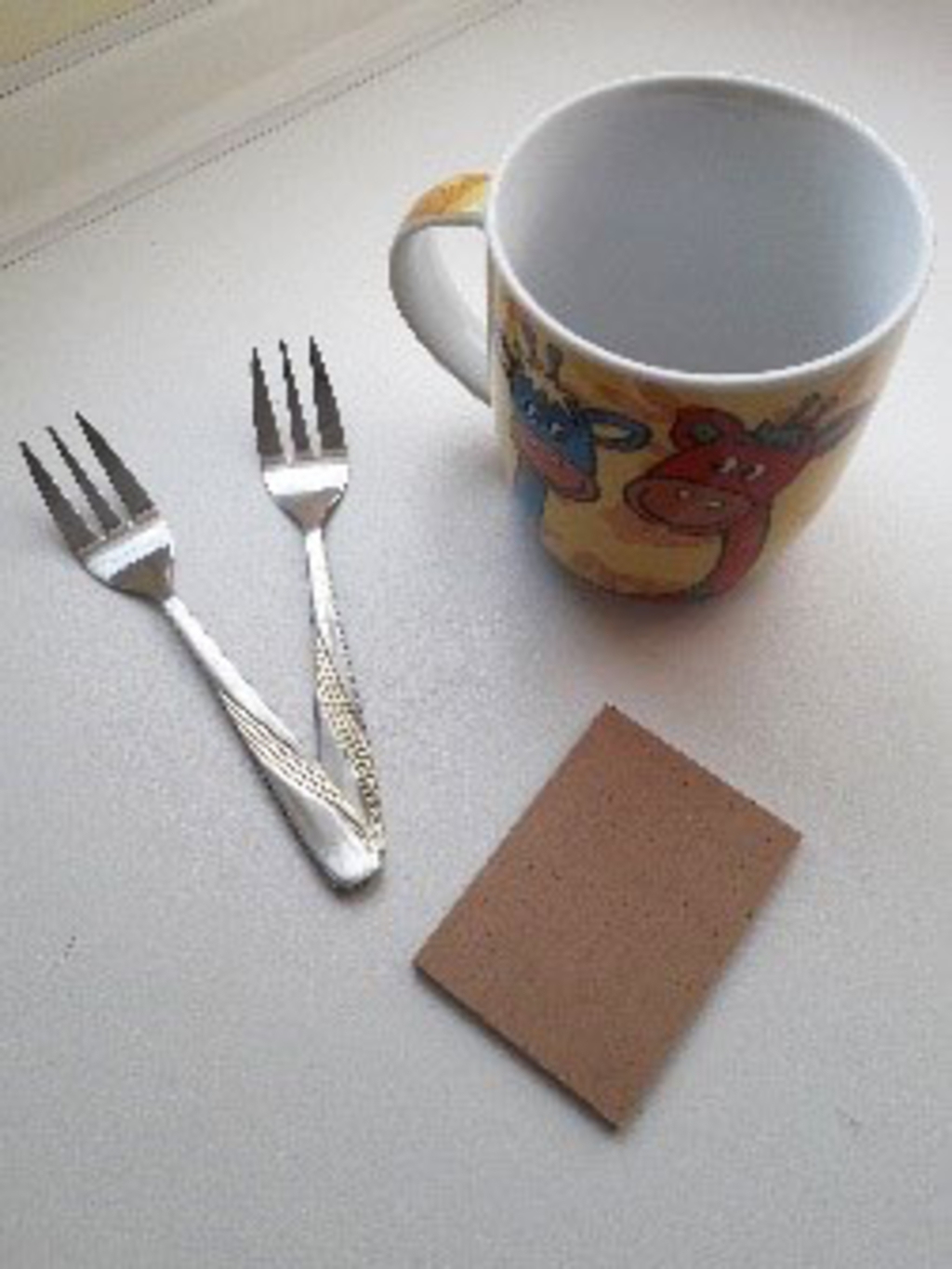 Das Bild zeigt eine bunte Tasse mit Tiermotiven, zwei kleine Gabeln und ein rechteckiges Stück Pappe. Diese Objekte könnten für verschiedene Bildungsaktivitäten oder Experimentierprojekte im schulischen Kontext verwendet werden.