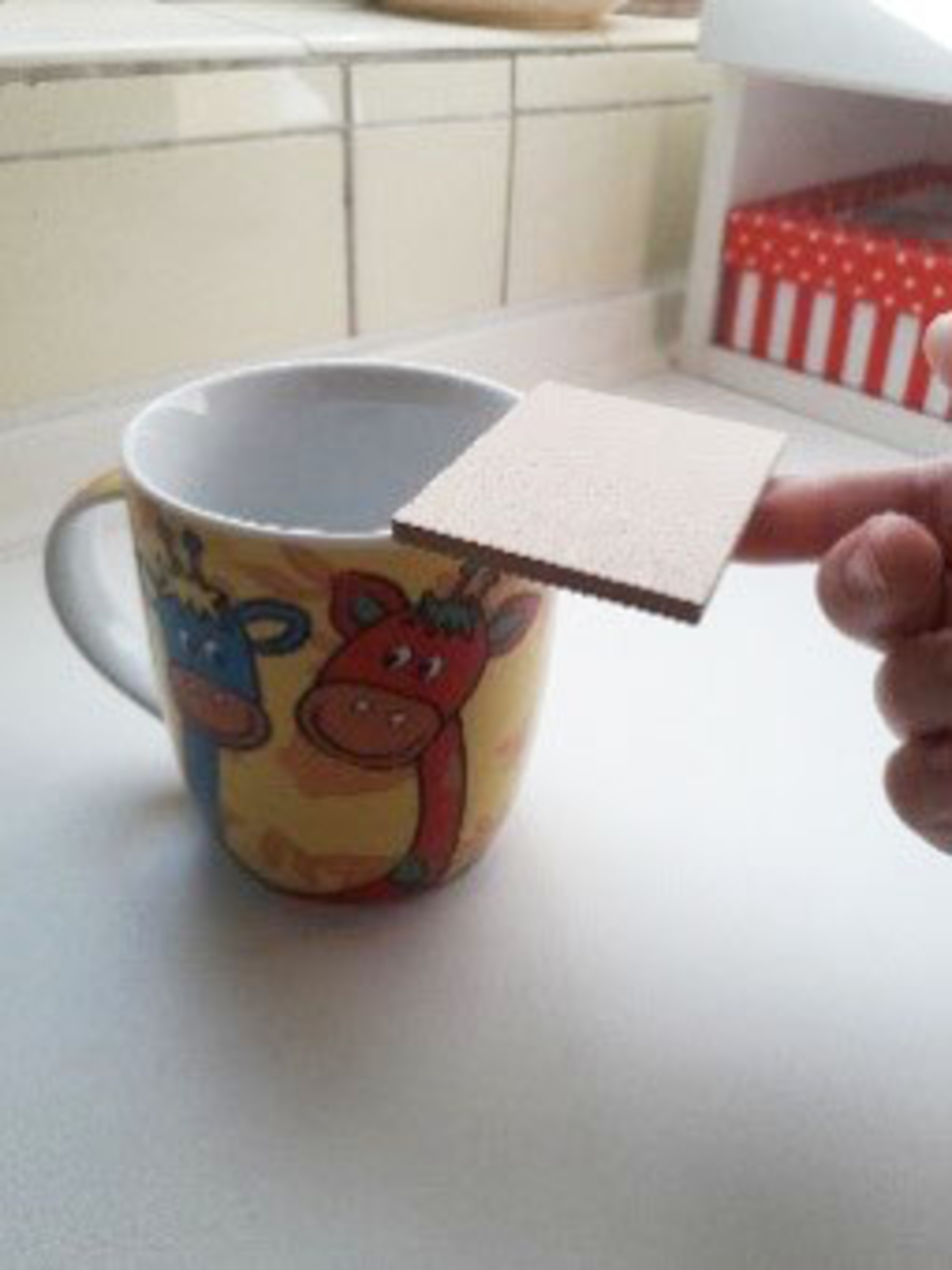 Das Bild zeigt eine bunte Tasse mit Tiermotiven, auf deren Rand eine kleine Pappe gelegt wird. Das ist Teil eines Experimentes zur Demonstration eines physikalischen Prinzips.