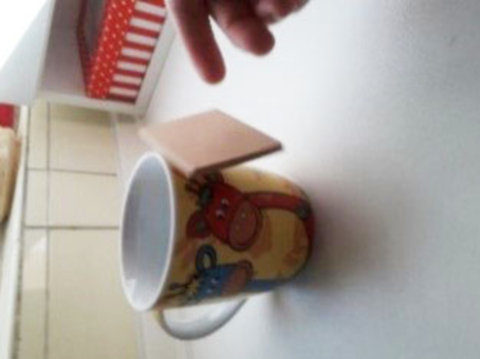 Das Bild zeigt eine bunte Tasse mit Tiermotiven, die auf einem Tisch steht. An der Tasse rechts befindet sich ein kleines Holzst&uuml;ck, das zu schweben scheint, daneben ist noch eine Hand zu erkennen, die das Holzst&uuml;ck nicht festh&auml;lt.