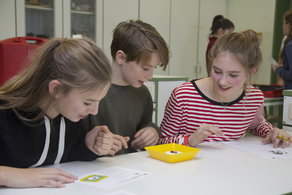 Auf dem Bild sind ein Schüler und zwei Schülerinnen zu sehen, die an einem Tisch sitzen und interessiert in einen gelben Behälter schauen, der einen selbstgebauten Kompass enthält. Sie scheinen sich aktiv auszutauschen, während sie möglicherweise Anweisun