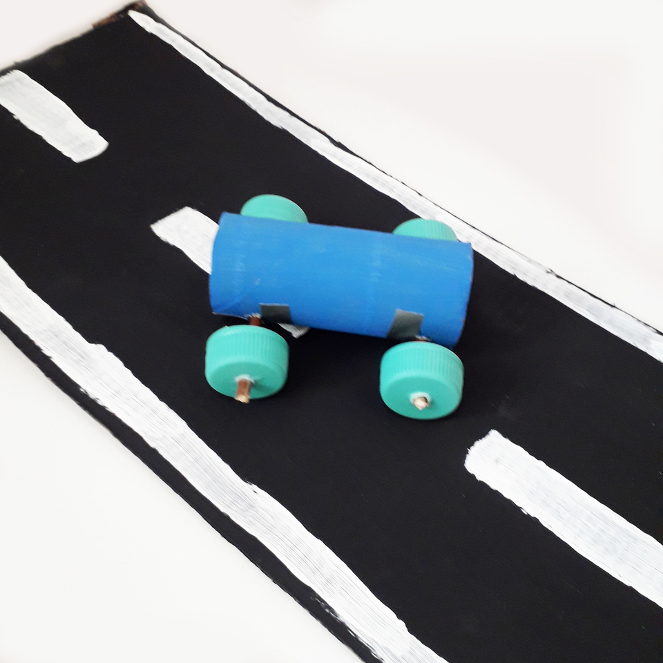 Das Bild zeigt ein selbstgebautes Fahrzeug aus recycelten Materialien, das auf einer kunstvoll gestalteten Stra&szlig;e platziert ist. Das Fahrzeug besteht aus einem blauen Zylinder mit gr&uuml;nen Radachsen, w&auml;hrend die Stra&szlig;e schwarz mit wei&szlig;en Linien dargestellt 