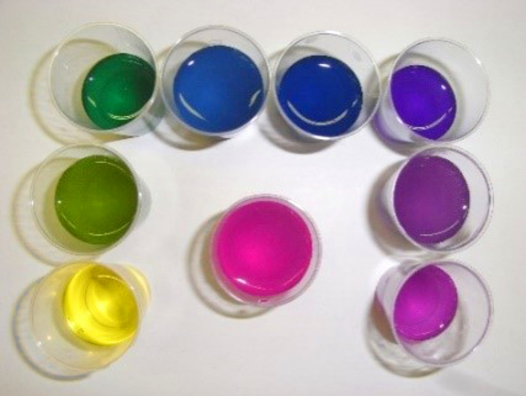 Das Bild zeigt eine Anordnung von neun durchsichtigen Bechern mit verschiedenen farbigen Flüssigkeiten. Diese Farben reichen von grün über blau und lila bis pink, und sind ideal für chemische Experimente im Bildungsbereich.