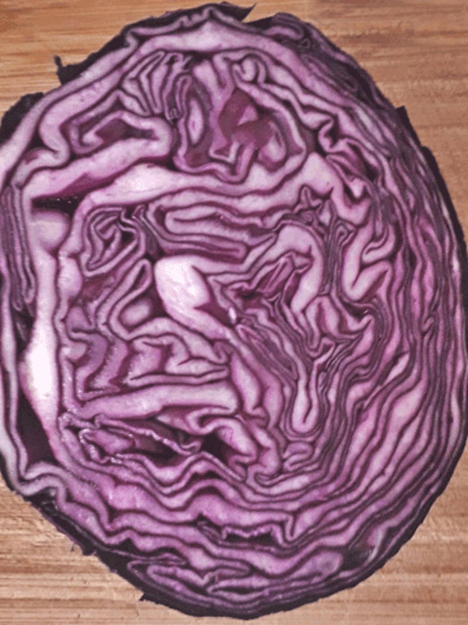 Das Bild zeigt einen Querschnitt von Rotkohl, der auf einem Holzuntergrund liegt. Die verschiedenen lila und wei&szlig;en Schichten des Kohls sind deutlich sichtbar und pr&auml;sentieren die charakteristische Struktur des Gem&uuml;ses.