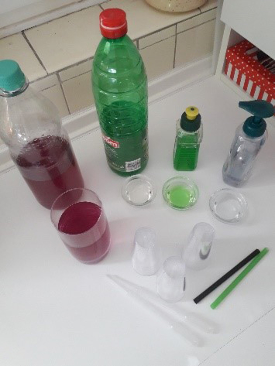 Das Bild zeigt eine Anordnung verschiedener Fl&uuml;ssigkeiten in Flaschen und Gl&auml;sern sowie einige Mess- und Experimentierger&auml;te, wie Pipetten und Bechergl&auml;ser. Die Farben der Fl&uuml;ssigkeiten reichen von Rot &uuml;ber Gr&uuml;n bis zu Transparent, passend f&uuml;r chemische E