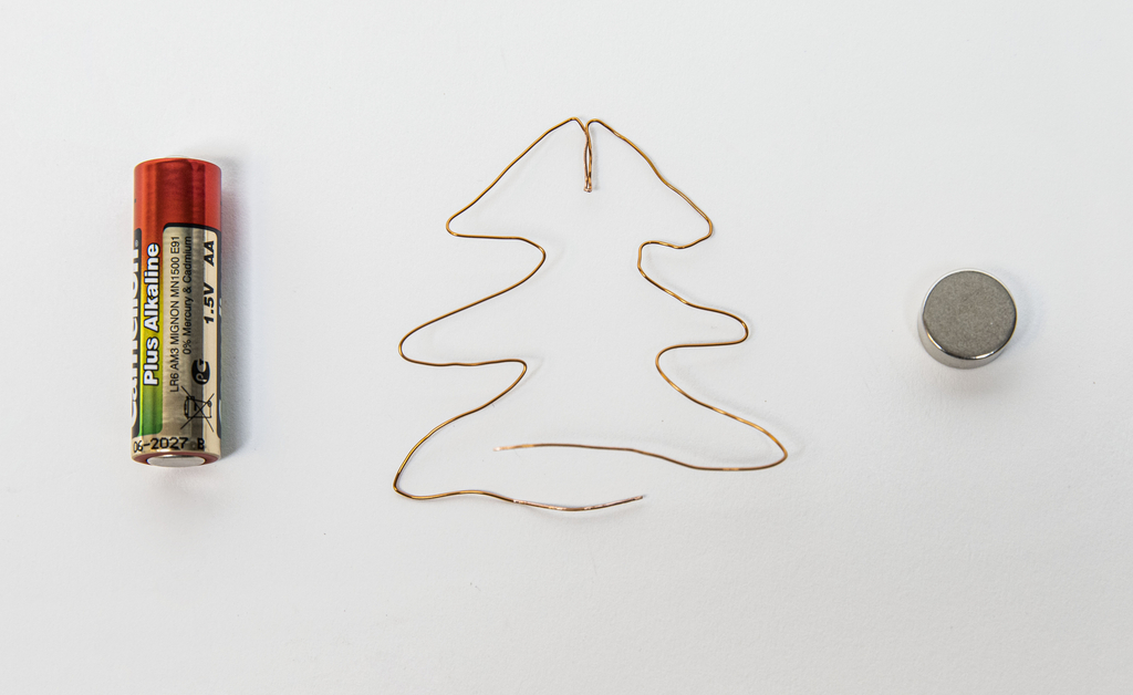 Das Bild zeigt eine AA-Batterie, einen kleinen Magneten und eine Drahtform in Form eines Weihnachtsbaums. Diese Materialien sind typisch für Experimente, die elektrische Grundlagen und Magnetismus veranschaulichen.