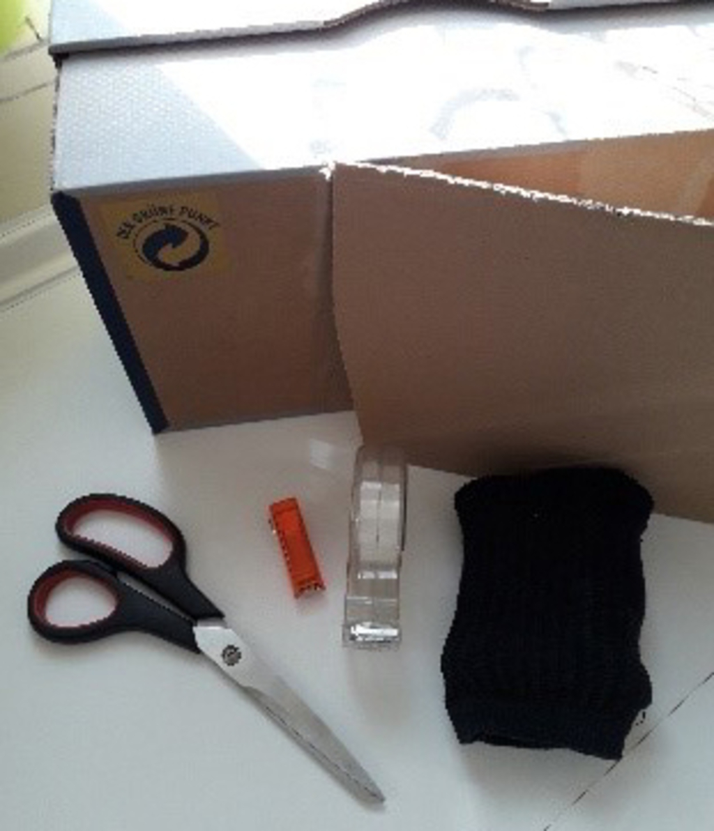 Das Bild zeigt eine Auswahl an Bastelmaterialien, die für Experimente genutzt werden können. Zu sehen sind eine Schere, Klebeband, eine Röllchenbatterie und ein schwarzer Stoff, daneben steht ein Karton.