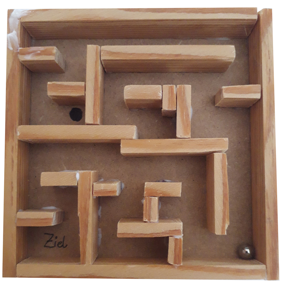 Das Bild zeigt ein einfaches selbstgebautes Labyrinth aus Holz mit verschiedenen St&auml;ben, durch die eine kleine Metallkugel hindurchrollen kann. Am unteren Ende ist ein markierter Zielpunkt zu erkennen, wo die Kugel ankommen soll.