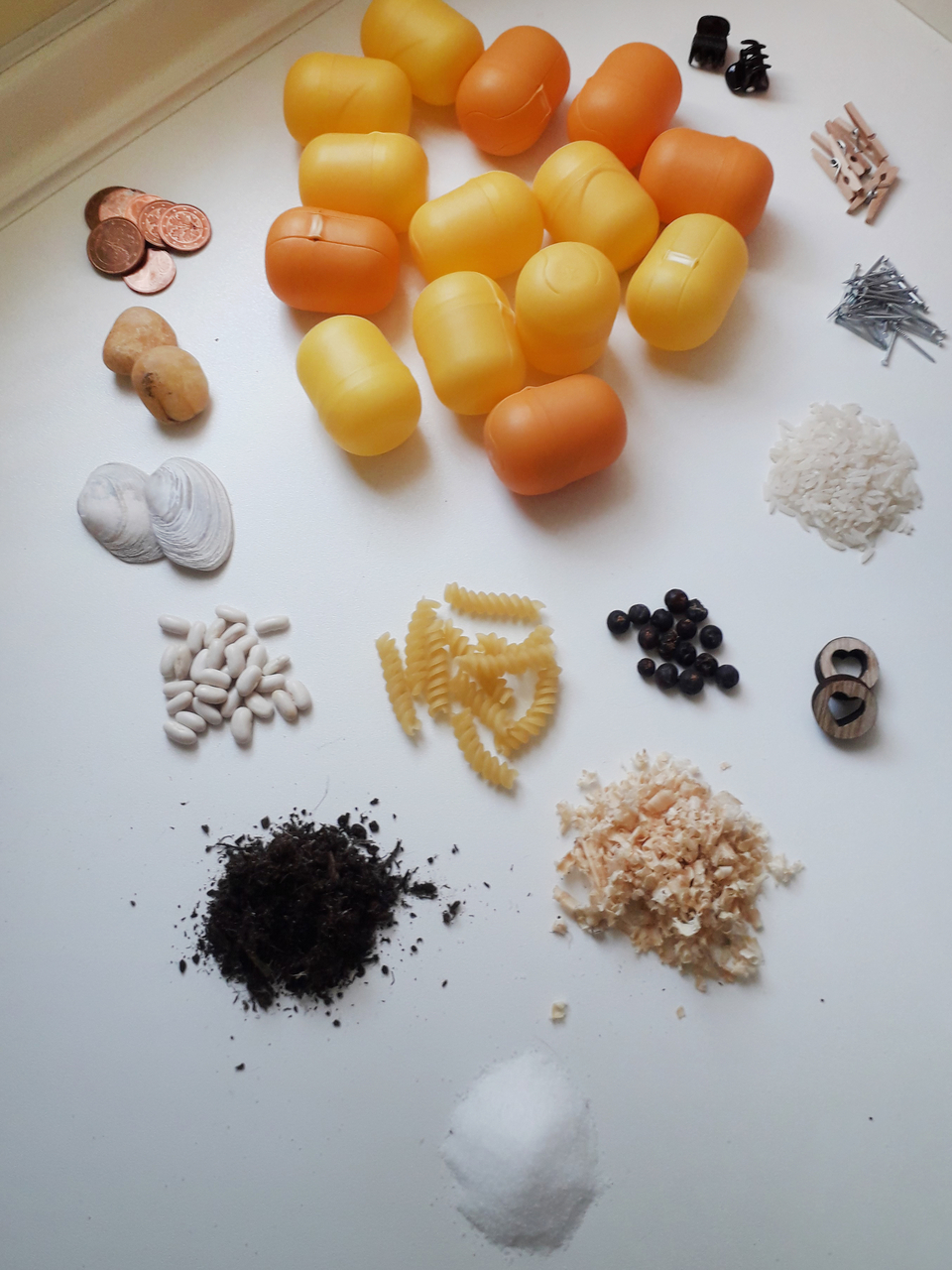 Das Bild zeigt eine Vielzahl von Materialien, die verwendet werden k&ouml;nnen, um ein H&ouml;rmemory zu bauen, darunter kleine bunte gelbe Plastikbeh&auml;lter, verschiedene Lebensmittel wie Reis und Pasta, sowie Gegenst&auml;nde wie M&uuml;nzen und N&auml;gel. Diese Materialien f&ouml;rd