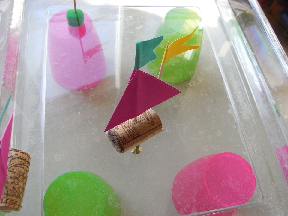 In einer klaren Wasserschale schwimmt ein kleiner Wasserfahrzeug, das aus einem Korken besteht und mit bunten Papierf&auml;hnchen dekoriert ist. Um den Korken herum sind mehrere farbige, halbtransparente Beh&auml;lter platziert, die in Pink und Gr&uuml;n gef&auml;rbt sind.