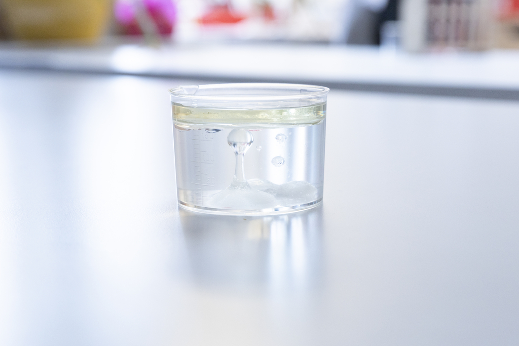 Das Bild zeigt ein Becherglas mit einer klaren Flüssigkeit und einer schaumigen Phase an der Unterseite. Es sind kleine Luftblasen zu sehen, die im Glas aufsteigen, während sich eine gelbliche Ölschicht an der Oberfläche bildet.