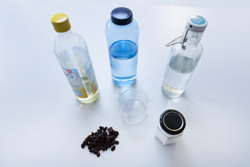 Das Bild zeigt mehrere Flaschen mit verschiedenen Fl&uuml;ssigkeiten, darunter &Ouml;l, Wasser und eine farblose Fl&uuml;ssigkeit, sowie einen Messbecher und eine kleine Dose. Auf dem Tisch liegen au&szlig;erdem eine Menge Rosinen, die vermutlich f&uuml;r ein Experiment verwendet 