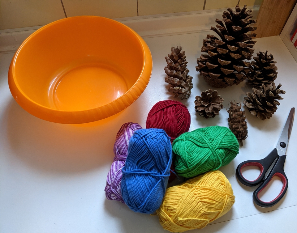 Ein orangefarbener Beh&auml;lter steht neben verschiedenen Kiefernzapfen und bunten Wollkn&auml;ueln in Blau, Gr&uuml;n, Rot, Lila und Gelb. Eine Schere liegt ebenfalls auf dem Tisch, was auf potenzielle Bastelaktivit&auml;ten hinweist.
