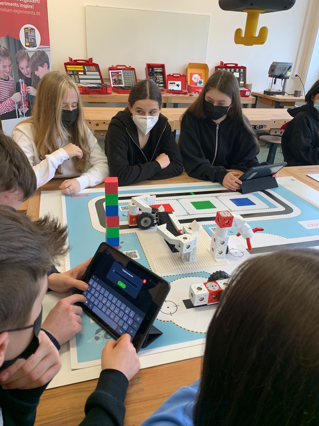 Das Bild zeigt eine Gruppe von Schülern, die an einem Tisch arbeiten, während sie Tablets zum Programmieren nutzen, um den eXperiBot Lernroboter zu steuern. Im Hintergrund sind verschiedene Experimentierkästen auf Regalen sichtbar, die auf eine Lernumgebu