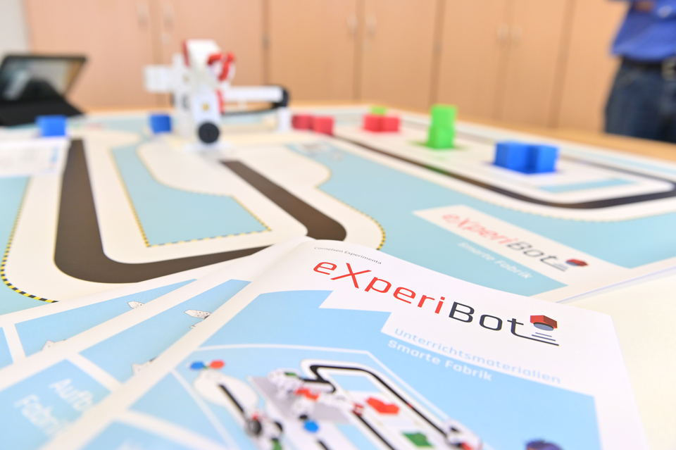 Das Bild zeigt einen Tisch mit einem Experimentier-Bereich, auf dem eine Strecke mit farbigen Bl&ouml;cken und einem kleinen Robotermodell angeordnet ist. Vorne liegen Unterrichtsmaterialien mit der Aufschrift "eXperiBot - Smarte Fabrik".