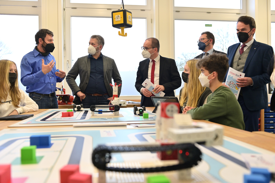 Das Bild zeigt eine Gruppe von Erwachsenen und Jugendlichen in einem Klassenraum, die an einem Tisch mit dem eXperiBot Lernroboter arbeiten. Ein Dozent erkl&auml;rt den Anwesenden etwas, w&auml;hrend mehrere Teilnehmer aufmerksam zuh&ouml;ren und sich Notizen machen.