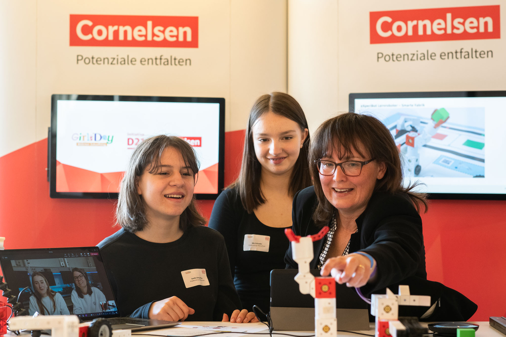 Das Bild zeigt drei Personen an einem Stand des Unternehmens Cornelsen, die sich mit dem eXperiBot Lernroboter besch&auml;ftigen. Im Hintergrund sind ein Laptop und Bildschirmanzeigen zu sehen, die auf Bildungsinitiativen hinweisen.