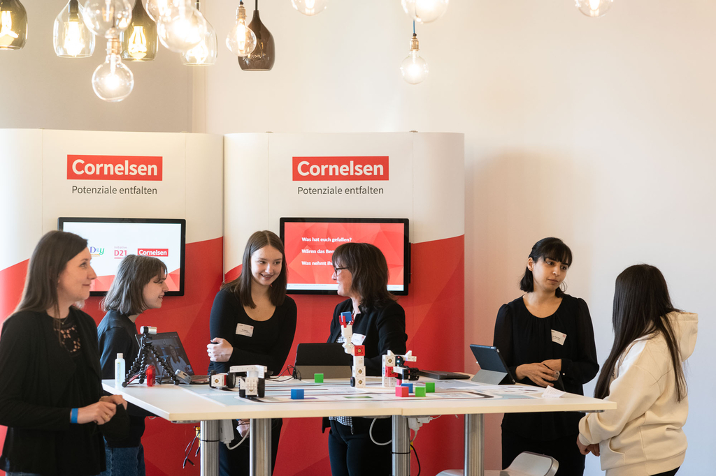 Das Bild zeigt einen Stand von Cornelsen, der auf Coding & Robotik für das Bildungswesen fokussiert ist. Mehrere Mädchen diskutieren und präsentieren Produkte vor einer modernen, ansprechenden Kulisse mit beleuchteten Lampen und Displays.