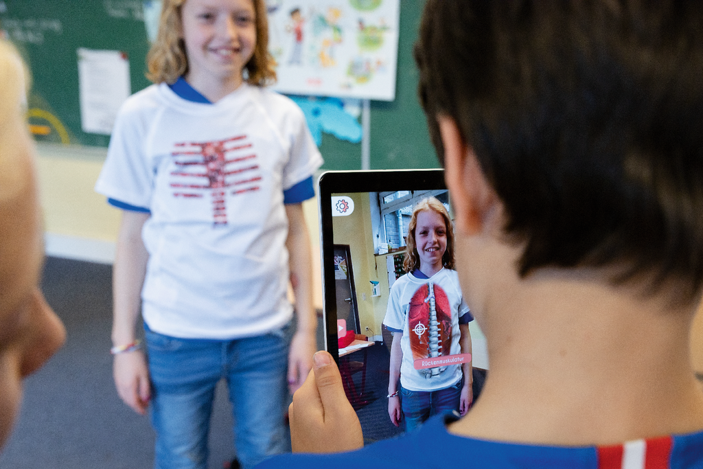 Das Bild zeigt ein Kind, das ein T-Shirt mit anatomischen Motiven tr&auml;gt und vor einer Tafel steht. Ein anderes Kind h&auml;lt ein Tablet, auf dem eine Augmented-Reality-Anwendung zu sehen ist, die die Anatomie des Tr&auml;gers darstellt.